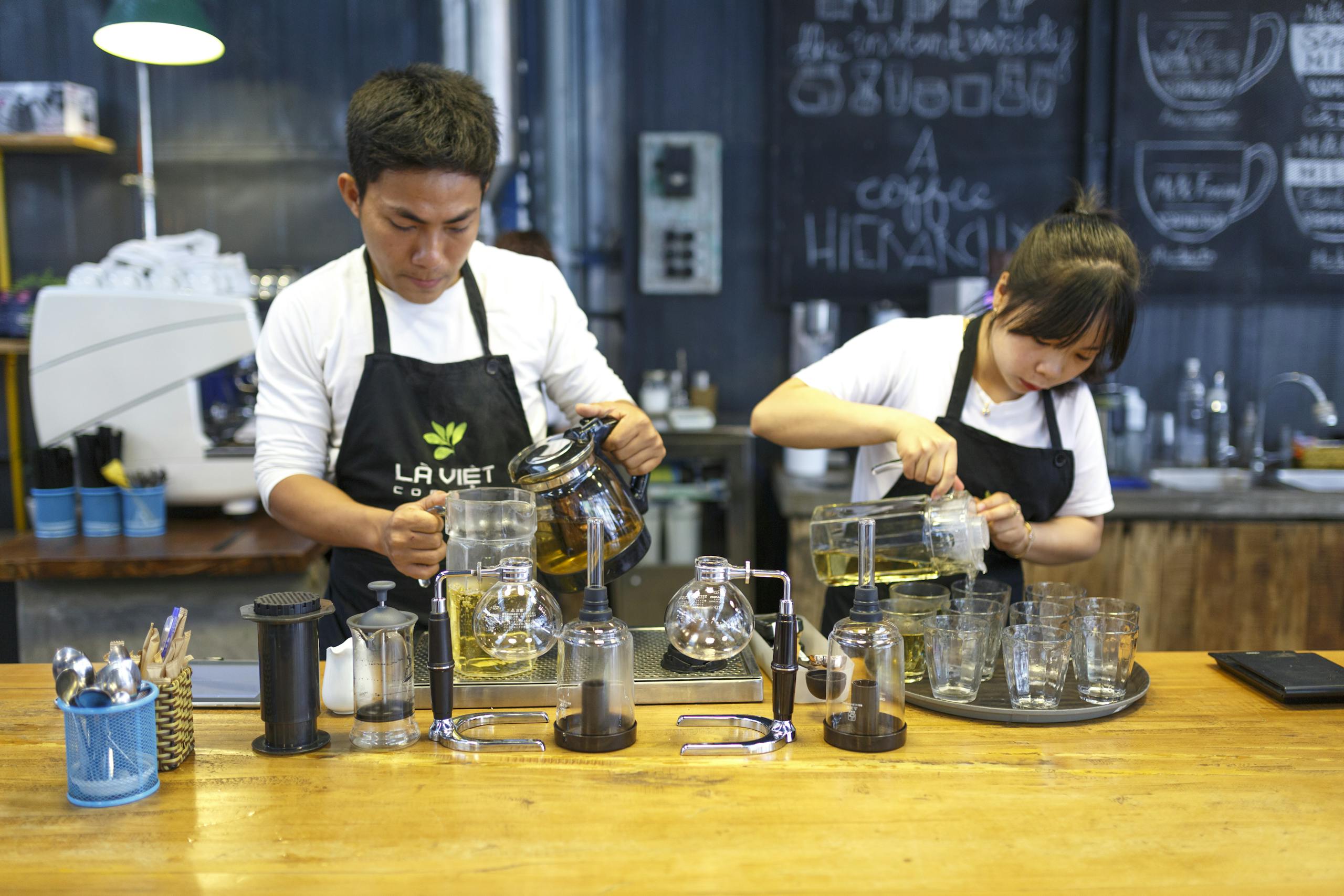 Two baristas are actively preparing beverages in a lively coffee shop setting.